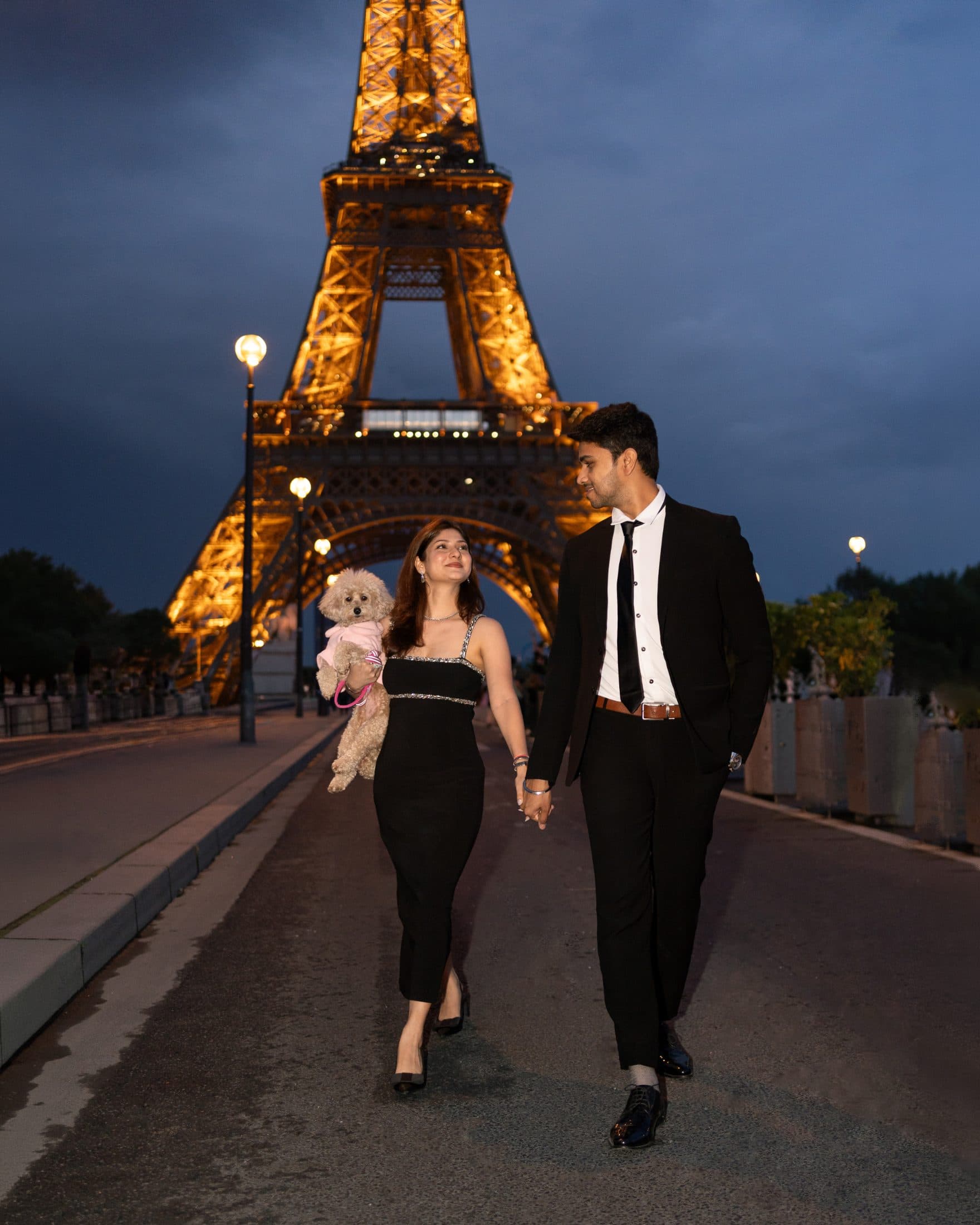 Couple walking with their dog at night near the illuminated Eiffel Tower during a romantic Paris couple photoshoot.